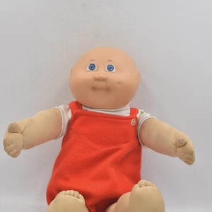 May include: A vintage Cabbage Patch Kid doll with a bald head, blue eyes, and a smiling expression. The doll is wearing a red jumpsuit over a white shirt. The doll is sitting with its arms and legs outstretched.