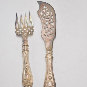 May include: A silver fork and knife set. The fork has four tines and an ornate handle. The knife has a decorative, openwork blade and a matching handle. The set is likely vintage and suitable for serving.