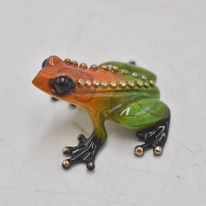 Puede incluir: Figurita de rana colorida con cabeza naranja, espalda verde y vientre amarillo. La rana tiene ojos negros y detalles dorados en la espalda y las patas. El objeto es decorativo.