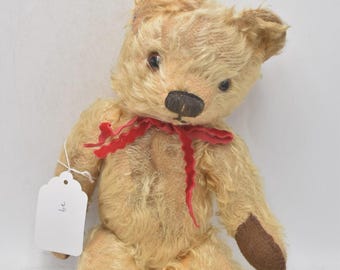 May include: A vintage teddy bear with light brown fur, a dark brown nose, and small black eyes. It has a red ribbon around its neck and brown felt paws. A white tag hangs from its arm.