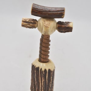 May include: A rustic bottle opener crafted from natural materials. It has a dark brown, textured base and a cream-coloured body with a wooden screw mechanism. The top has a cross-shaped design with brown antler pieces.