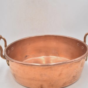May include: A copper bowl with two curved handles. The bowl has a wide, shallow design and a polished finish. The copper has a warm, reddish-brown hue. The handles are attached to the sides of the bowl.