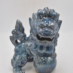 May include: A teal-blue glazed ceramic Foo Dog statue. The mythical creature has a detailed mane, a smiling expression, and holds a ball. This decorative statue is a traditional Asian art piece.