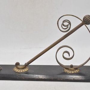 May include: An antique wall-mounted coat rack. It features a dark wooden base with a decorative metal design. The metalwork includes a hook, a rod, and ornate swirls, with brass-colored accents. The rack is designed for hanging items.