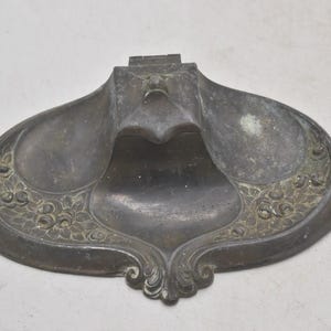 May include: Antique metal inkwell with a decorative floral design. The inkwell has a dark, aged appearance with a central ink reservoir and two side wells. The edges are adorned with embossed floral patterns, and the overall shape is ornate.