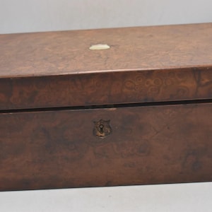 May include: A rectangular wooden box with a dark brown finish and a decorative burl wood pattern. The box has a small, light-coloured accent on the lid and a keyhole on the front. The box is likely used for storage.