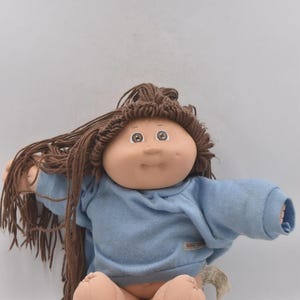 May include: A vintage Cabbage Patch Kid doll with brown yarn hair and a light blue sweater. The doll has a round face, rosy cheeks, and brown eyes. The doll is sitting with its arms outstretched.