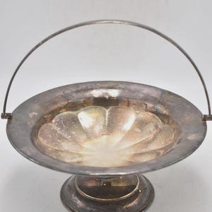 May include: A silver-toned, footed serving dish with a handle. The dish has a scalloped interior design and a circular base. The handle arches over the dish. The metal has a tarnished appearance, suggesting age.
