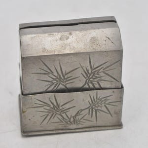 May include: A small, silver-coloured box with a hinged lid. The box is rectangular with rounded edges and features etched bamboo leaf designs on the front and top. The box is made of metal and has a slightly aged appearance.