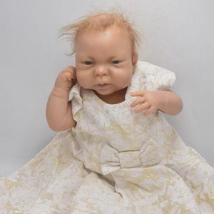 May include: A lifelike baby doll with fair skin and light brown hair. The doll wears a white dress with a gold floral pattern and a bow at the waist. The doll's expression is serious, with a slight frown.