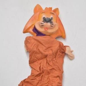 May include: A vintage lion puppet with an orange head and body. The lion's face has black whiskers, a black nose, and a red mouth. The puppet's body is made of orange fabric, and it has small tan hands.