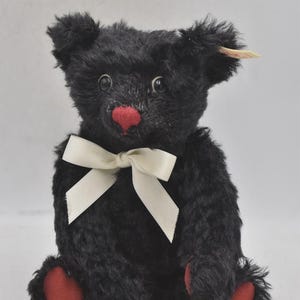 May include: A black teddy bear with a red nose and feet, and a white bow. The bear has a tag on its ear. The bear is a classic toy.