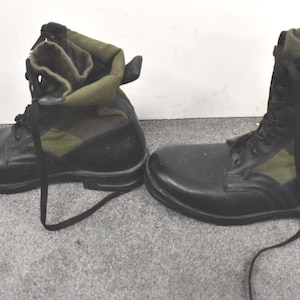 May include: A pair of black combat boots with olive green canvas uppers. The boots feature black laces and a robust sole. These boots are designed for durability and are suitable for outdoor use.