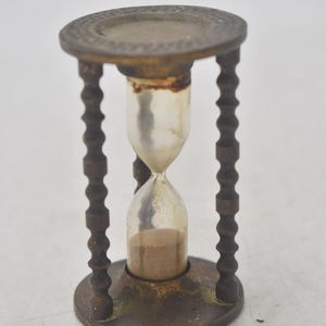 Op de afbeelding: Een antieke zandloper met een heldere glazen bol met zand, ondersteund door een donkerbruin, sierlijk metalen frame. De zandloper heeft een decoratieve boven- en onderkant en de poten zijn ingewikkeld gesneden.
