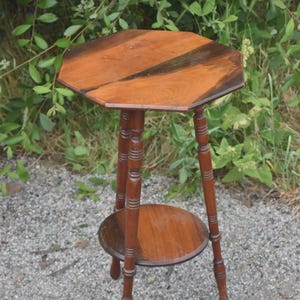 May include: A wooden side table featuring an octagonal top with a two-tone brown wood grain pattern. The table stands on three slender, turned legs that are connected by a circular lower shelf, providing a stable structure for this piece of furniture.