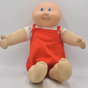 May include: A vintage Cabbage Patch Kid doll with a bald head, blue eyes, and a smiling expression. The doll is wearing a white shirt and a red overall. The doll is sitting with its arms and legs outstretched.