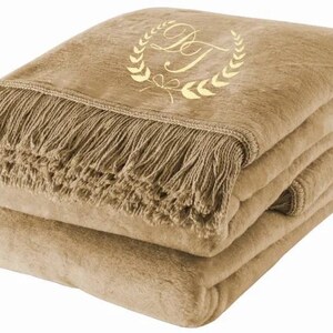 Può includere: Una coperta in pile marrone con un bordo a frangia e un monogramma ricamato in oro in un design a corona.