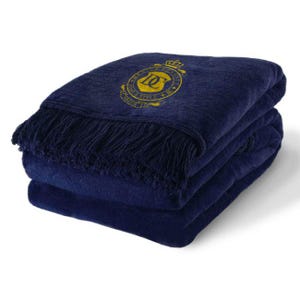 Pode incluir: Uma manta de arremesso azul marinho com um brasão bordado em ouro que apresenta uma coroa, um escudo com a letra "D" e as palavras "DOMINUS PROVIDEBIT". A manta tem uma borda de franjas.