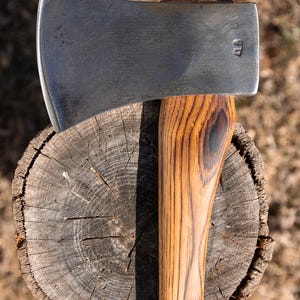 May include: Axe with a wooden handle and a steel blade, resting on a tree stump. The axe head is silver and the handle is a light brown color. The axe is a tool for chopping wood.