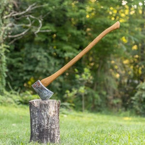May include: A felling axe with a long, curved wooden handle and a metallic head featuring a weathered, dark grey finish. The axe is embedded into the top of a cylindrical, rough-textured wooden log stump, standing upright in an outdoor setting.