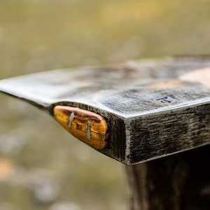 May include: A hand-forged steel axe head with a textured, dark metallic finish and a light-colored wooden handle. The axe head features a distinct wooden wedge insert secured with two metal pins, showing a weathered, industrial appearance.