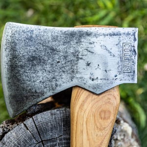 May include: A metal axe head with a weathered, dark gray patina and the word COLLINS stamped into the side. The axe head is attached to a light brown wooden handle, showing a clear wood grain pattern throughout the surface of the handle.