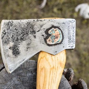May include: A metal axe head with a weathered, dark-spotted finish attached to a light-colored wooden handle. The metal surface features the stamped text COLLINS and 1 1/4, along with a circular logo containing the word Collins in blue script.