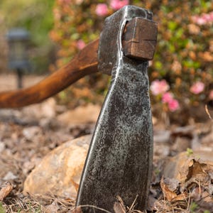 May include: A vintage axe with a weathered, dark metal head and a wooden handle. The axe is positioned in a bed of dry grass and leaves, with a blurred background of greenery and pink flowers.