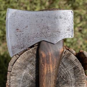 May include: A hand axe featuring a weathered, grey steel head with visible surface oxidation and a dark, stained wooden handle. The axe head is securely attached to the handle, showcasing a curved cutting edge and a textured metal finish.