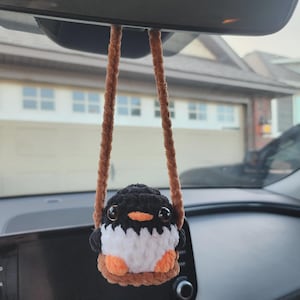 May include: A handmade crochet penguin car charm. The penguin has a black head, white chest, and orange feet and beak. It hangs from a brown cord, suspended from a car's rearview mirror. The charm is a cute, decorative accessory.
