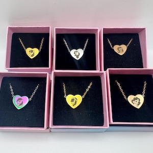 May include: Heart-shaped pendant necklaces in gold, silver, and iridescent finishes, each featuring a character portrait. The necklaces are displayed in pink boxes with black interiors, showcasing the jewelry's design and presentation.