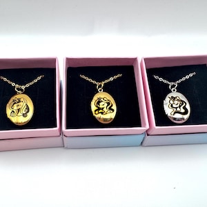 May include: Three oval-shaped locket necklaces in pink boxes. Two are gold-toned, one is silver-toned. Each locket features a black silhouette design of a character on a delicate chain. The lockets are displayed on a black background.