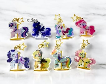 May include: Eight colorful pony-themed keychains with gold star accents. Each keychain features a different pony design with vibrant colors and intricate details. The keychains are attached to gold-colored metal rings.