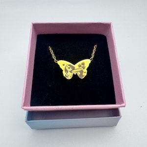 May include: A gold-colored butterfly pendant necklace displayed in a pink and blue jewelry box. The butterfly pendant has a detailed design. The necklace is on a black velvet background.