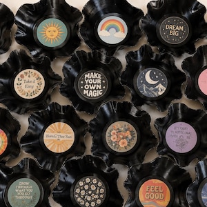 May include: A collection of black vinyl record bowls with colorful, circular designs in the center. Each bowl features a different design, including a rainbow, sun, moon, and various inspirational phrases like "Good Vibes" and "Dream Big".