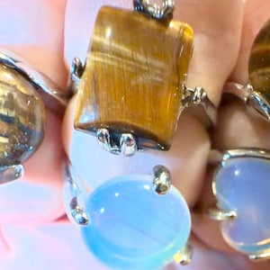 Puede incluir: Una colección de anillos de piedras preciosas con bandas plateadas. Los anillos presentan varias piedras, incluyendo ojo de tigre y una piedra azul claro. Las piedras de ojo de tigre tienen patrones marrones y dorados. Un anillo tiene una piedra rectangular, otros son ovalados o en forma de corazón.