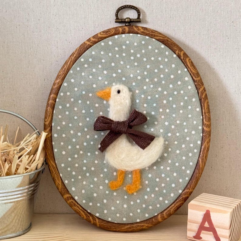 May include: An oval-shaped, framed wall hanging featuring a felted white goose with an orange beak and feet, and a brown bow. The goose is set against a light blue background with white polka dots. The frame is made of wood.