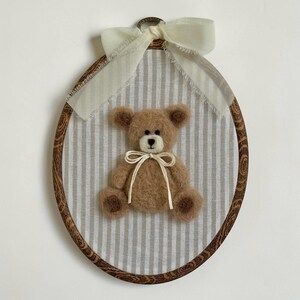 May include: An oval embroidery hoop frame with a dark wood grain pattern containing a light beige and white striped fabric. A fuzzy brown needle-felted teddy bear with a cream-colored muzzle is centered, adorned with a thin cream ribbon bow.