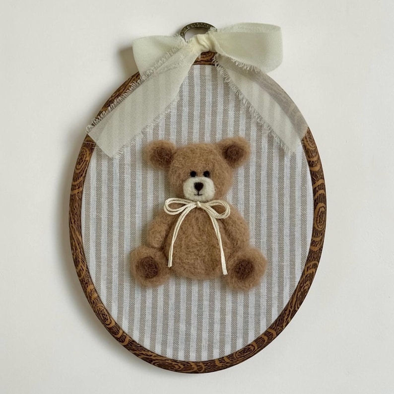 May include: An oval embroidery hoop frame with a dark wood grain pattern containing a light beige and white striped fabric. A fuzzy brown needle-felted teddy bear with a cream-colored muzzle is centered, adorned with a thin cream ribbon bow.