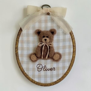 May include: An oval embroidery hoop frame containing a beige and white gingham fabric background. A brown fuzzy teddy bear with a tan ribbon bow around its neck is centered. The name Oliver is embroidered in brown thread below the bear.