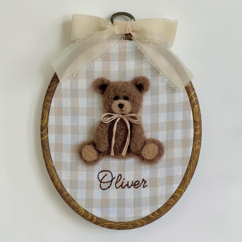 May include: An oval embroidery hoop frame containing a beige and white gingham fabric background. A brown fuzzy teddy bear with a tan ribbon bow around its neck is centered. The name Oliver is embroidered in brown thread below the bear.