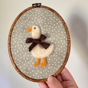 May include: A handmade felt duckling embroidery in a wooden hoop. The duckling is white with an orange beak and feet, and a brown bow. The background is gray fabric with white polka dots. The hoop is approximately 6 inches in diameter.