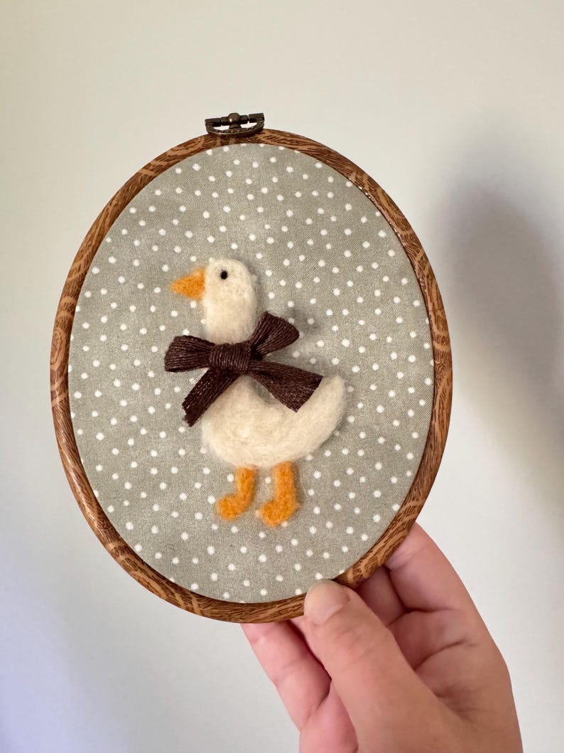 May include: A handmade felt duckling embroidery in a wooden hoop. The duckling is white with an orange beak and feet, and a brown bow. The background is gray fabric with white polka dots. The hoop is approximately 6 inches in diameter.