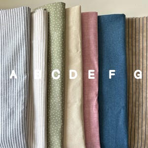 Seven neatly arranged fabric swatches labeled A to G, featuring stripes, polka dots, and solid colors in soft tones like gray, beige, olive, pink, blue, and rustic brown — fabric options for handmade textile art
