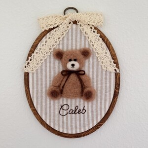 May include: An oval wooden embroidery hoop frame containing a light brown needle-felted teddy bear with a dark brown ribbon tied around its neck. The bear is centered on a beige and white striped fabric with the name Caleb embroidered in brown.