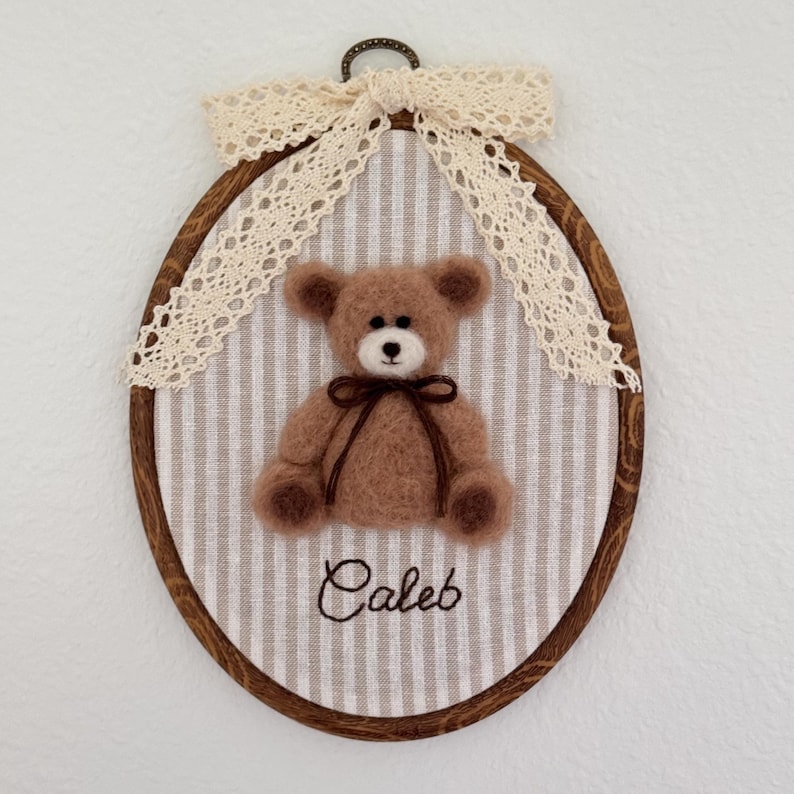 May include: An oval wooden embroidery hoop frame containing a light brown needle-felted teddy bear with a dark brown ribbon tied around its neck. The bear is centered on a beige and white striped fabric with the name Caleb embroidered in brown.