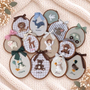 May include: A collection of circular wooden embroidery hoops featuring felt animal and character appliqués on fabric backgrounds. Each hoop is decorated with a fabric bow and includes embroidered names such as Noah, Emma, Grace, Charlie, Jack, Lucas, and James.