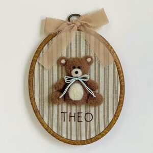 May include: An oval wooden embroidery hoop frame containing a beige and white striped fabric background. A brown felted teddy bear with a cream belly and light blue ribbon sits in the center. The name THEO is embroidered below in brown thread.