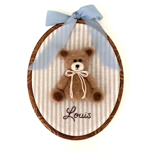 May include: An oval embroidery hoop frame containing a light brown felted teddy bear centered on a white and gray striped fabric background. The bear wears a thin cream ribbon bow. Below the bear, the name Louis is embroidered in dark brown thread.