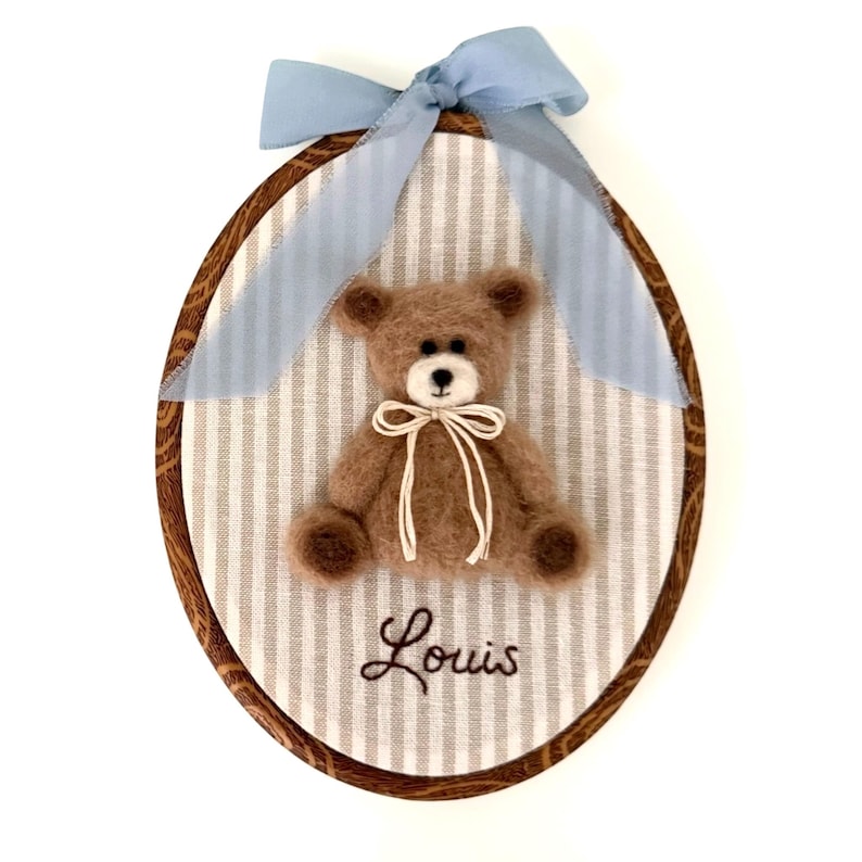 May include: An oval embroidery hoop frame containing a light brown felted teddy bear centered on a white and gray striped fabric background. The bear wears a thin cream ribbon bow. Below the bear, the name Louis is embroidered in dark brown thread.